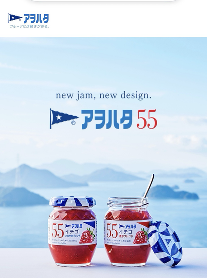 Two Aohata 55 strawberry jam jars with blue and white patterned lids. One jar is open, with a spoon inside. A blue sea and mountains are in the background.