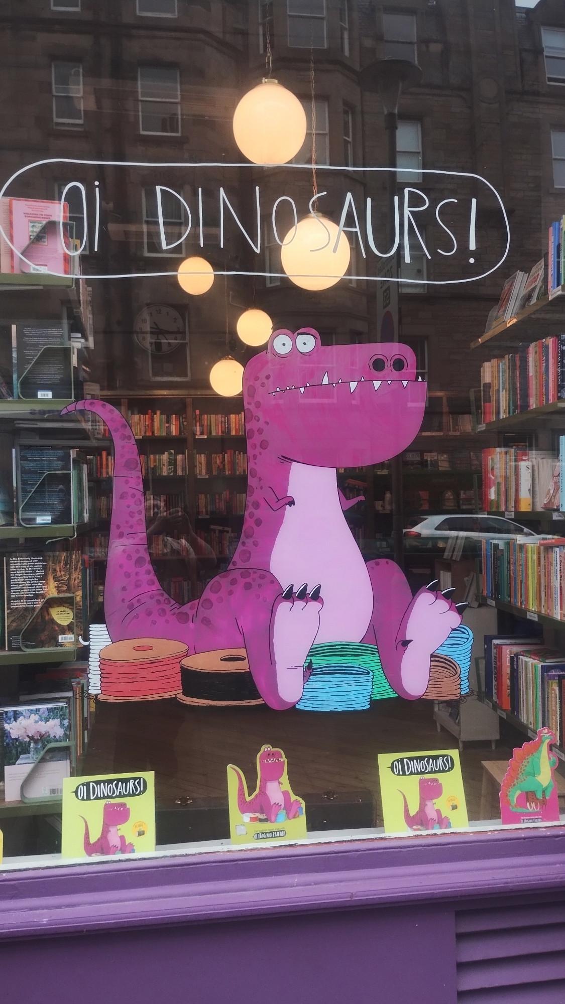 Finished window for Oi Dinosaurs, viewed from the outside, our large, purple dino sitting and looking out the glass