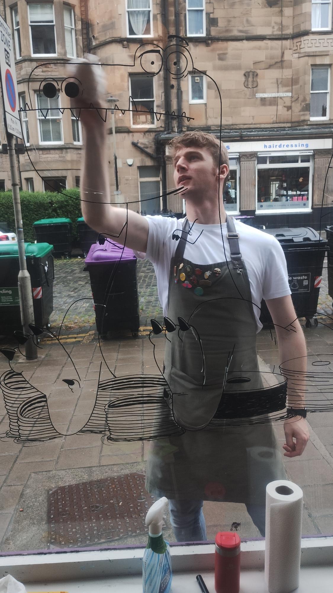 Looking out the bookshop window at bookseller Jack as he draws an outline for the window painting