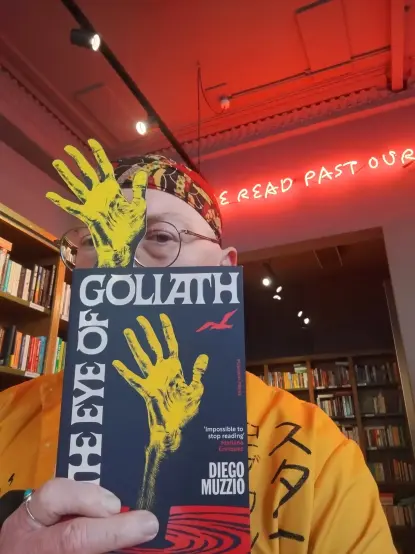 Bookseller Joe holding up a paperbackk copy of The Eye of Goliath, with a long, yellow, hand and arm bookmark design (imitating the book's cover) stickig out of the top, in the Edinburgh Bookshop