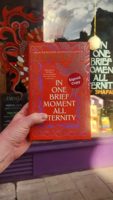 holding up a signed hardback copy of In One Brief Moment All Eternity, outside the window painted in its honour, at the Edinburgh Bookshop