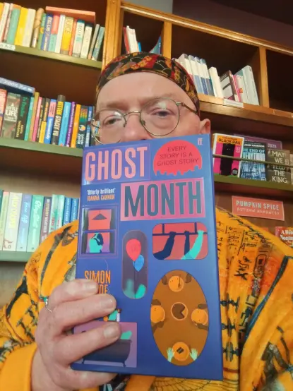 Bookseller Joe holding up a blue hardback edition of Ghost Month, by Simon Wroe, in front of the bookshelves in the Edinburgh Bookshop